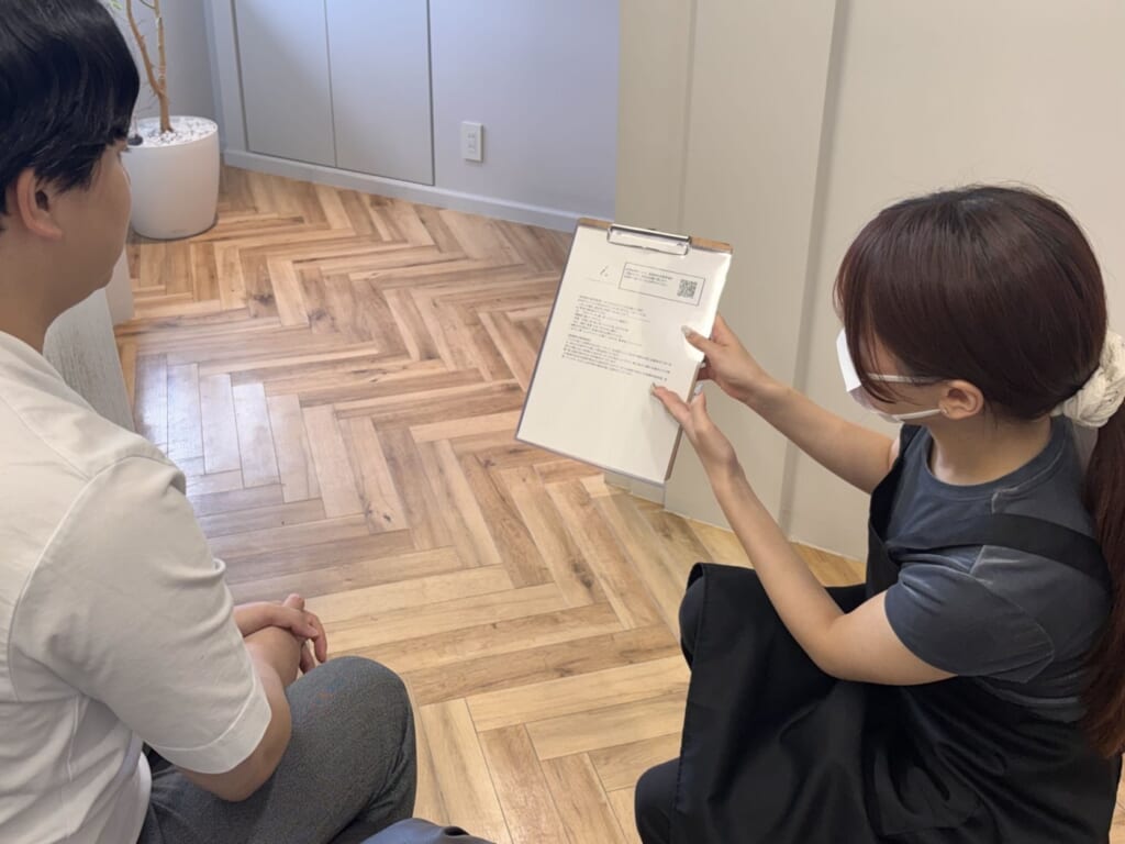 施術ルームで、スタッフがお客様に紙の資料を見せながら説明している様子